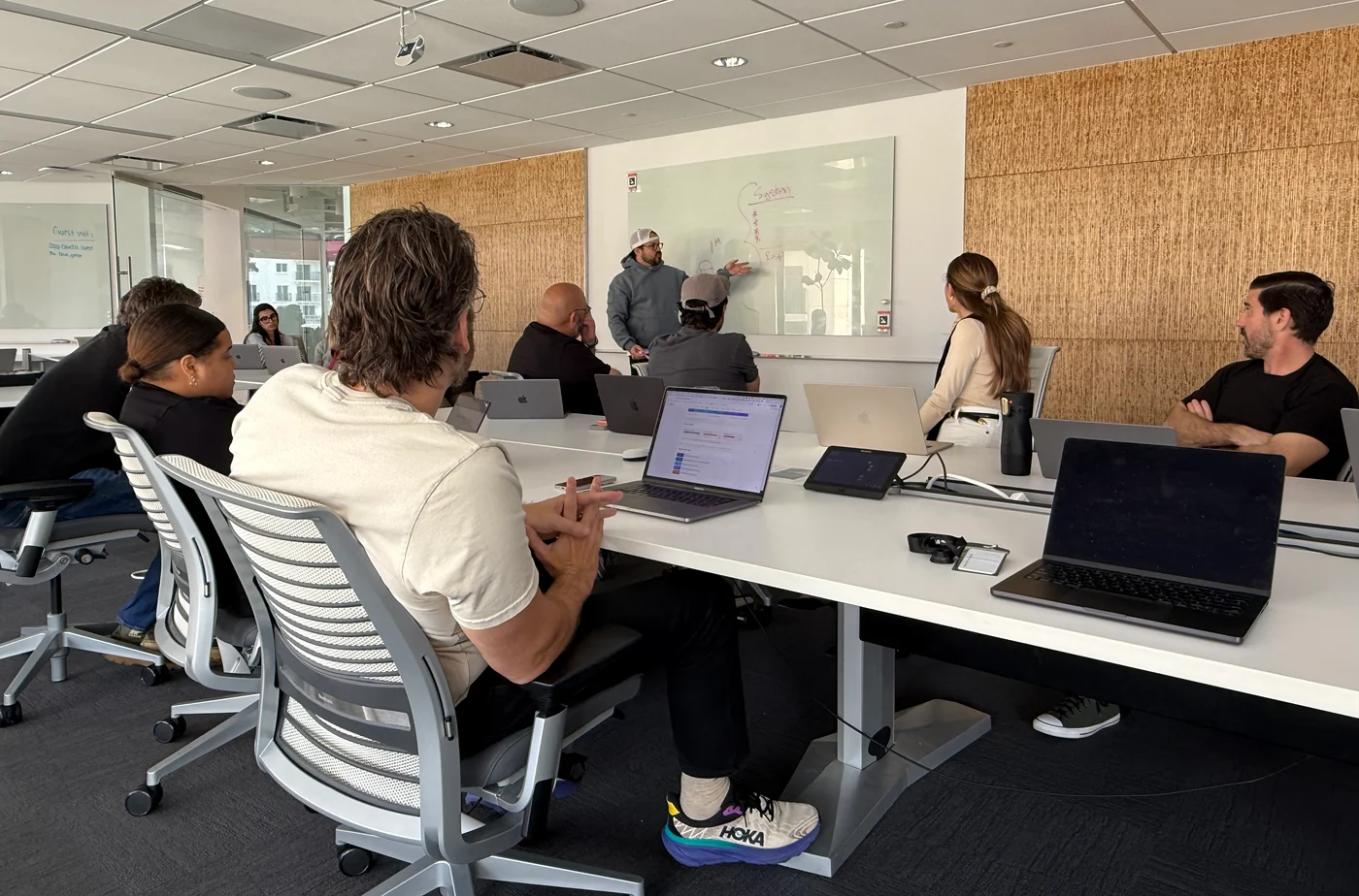 A presenter at a whiteboard with people seated around laptops.